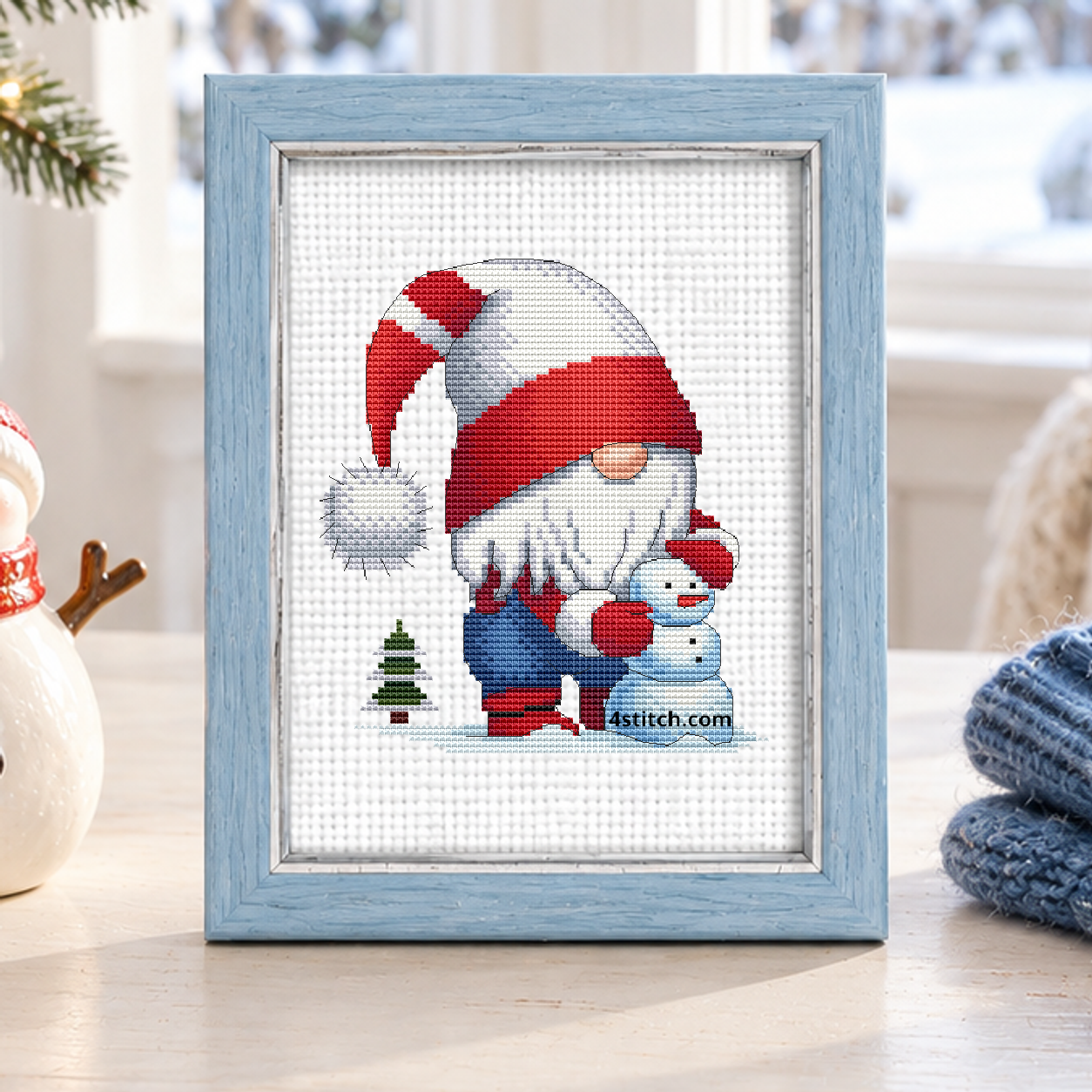 Snowman Christmas Gnome with a gnome in a red-and-white striped hat hugging a pale blue snowman beside a tiny fir tree, on white Aida in a powder-blue frame in a bright winter sunroom.