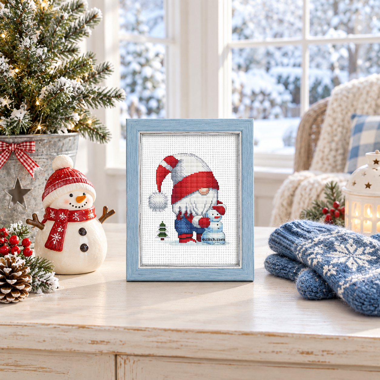 Snowman Christmas Gnome with a gnome in a red-and-white striped hat hugging a pale blue snowman beside a tiny fir tree, on white Aida in a powder-blue frame in a bright winter sunroom.
