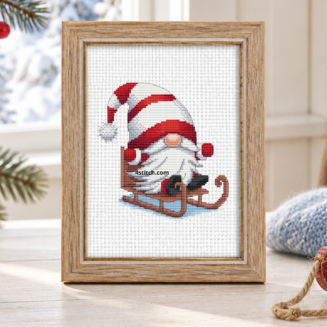 Sledding Christmas Gnome with a red-and-white striped-hat gnome riding a wooden sled across pale blue snow, on white Aida in a pale oak frame in a bright Scandinavian winter cabin.