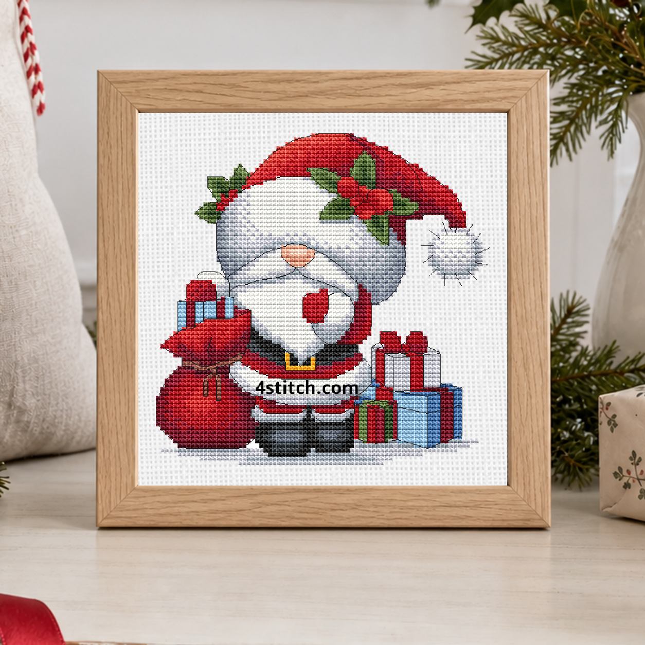 Santa Gifts Gnome with a holly-trimmed red Santa hat, a red gift sack and stacked Christmas presents, on white Aida in a pale oak frame on a bright Christmas gift-wrapping console.