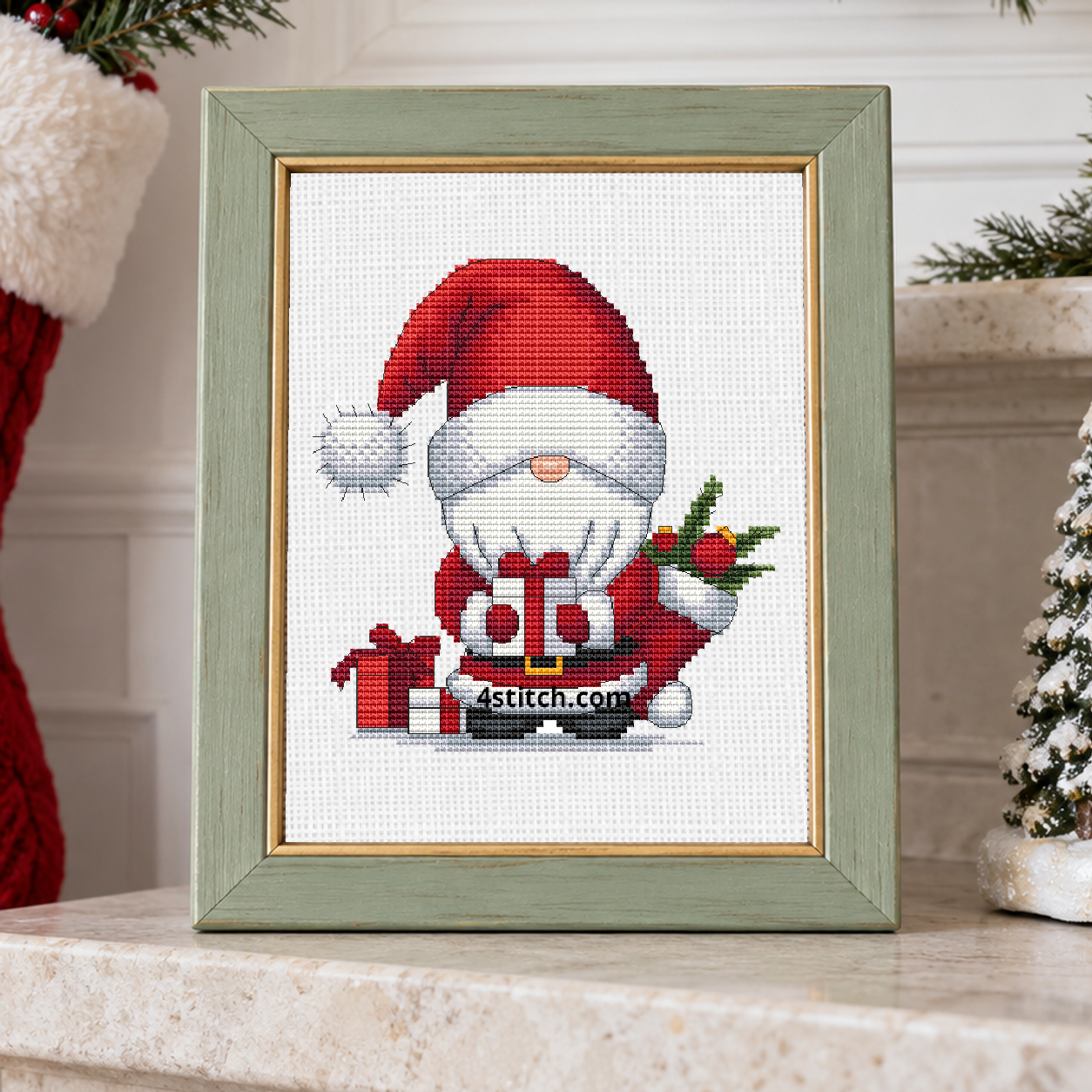 Santa Stocking Gifts Gnome with a red Santa hat, a small wrapped present and a Christmas stocking filled with greenery, on white Aida in a sage wooden frame on a light Christmas mantel.