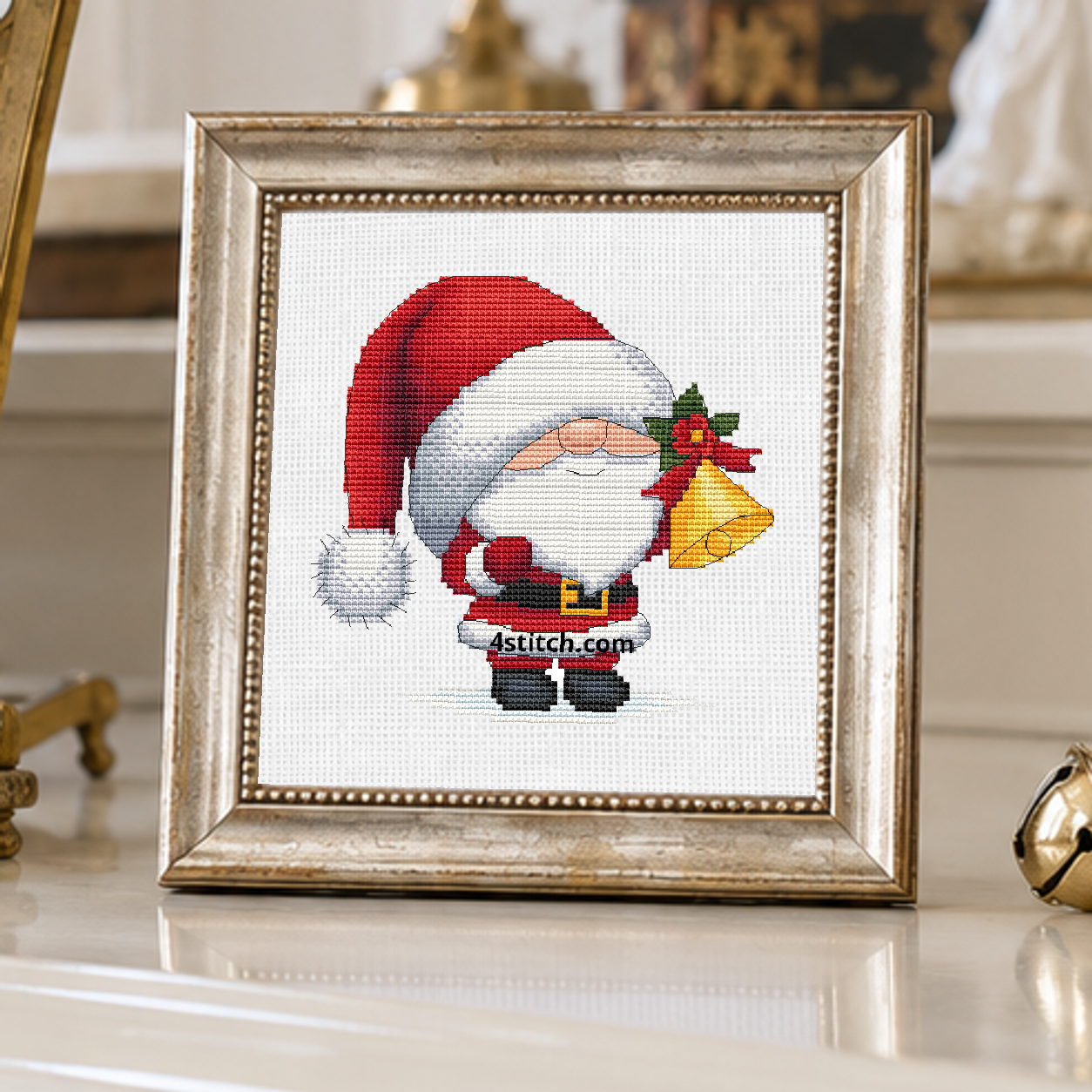 Jingle Bell Santa Gnome with a drooping red Santa hat and a golden handbell decorated with holly, on white Aida in a champagne frame beside festive bells in a sunlit room.