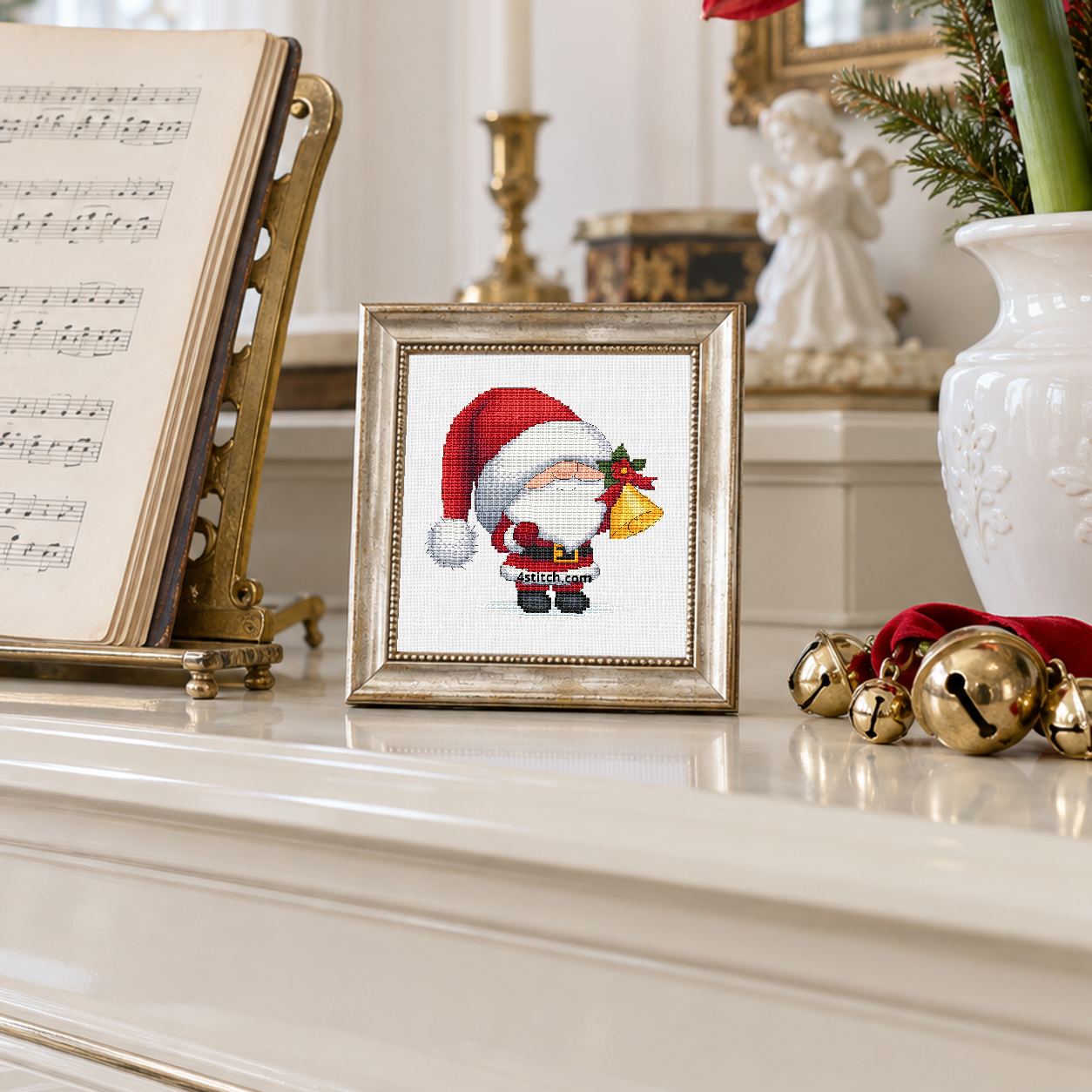 Jingle Bell Santa Gnome with a drooping red Santa hat and a golden handbell decorated with holly, on white Aida in a champagne frame beside festive bells in a sunlit room.