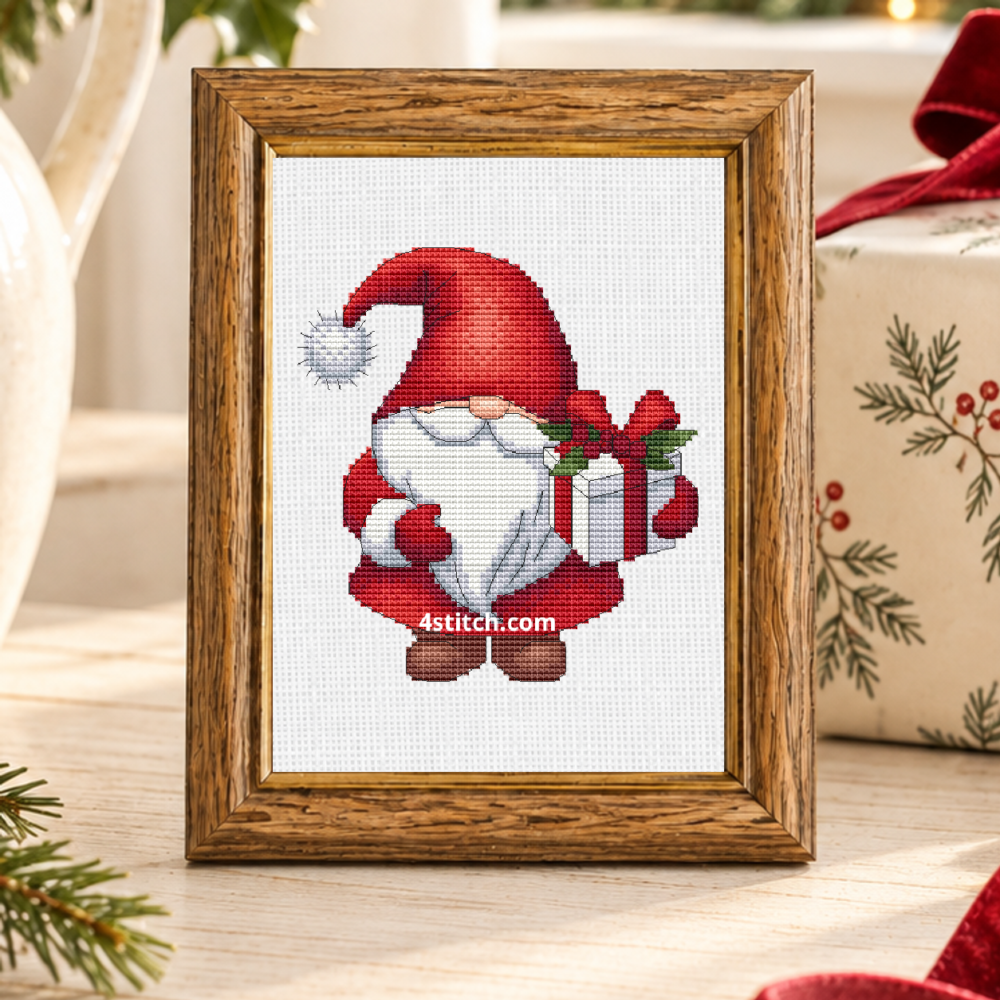 Close-up of Holly Gift Santa Gnome on white Aida in a honey-oak frame on a sunlit Christmas gift-wrapping console.