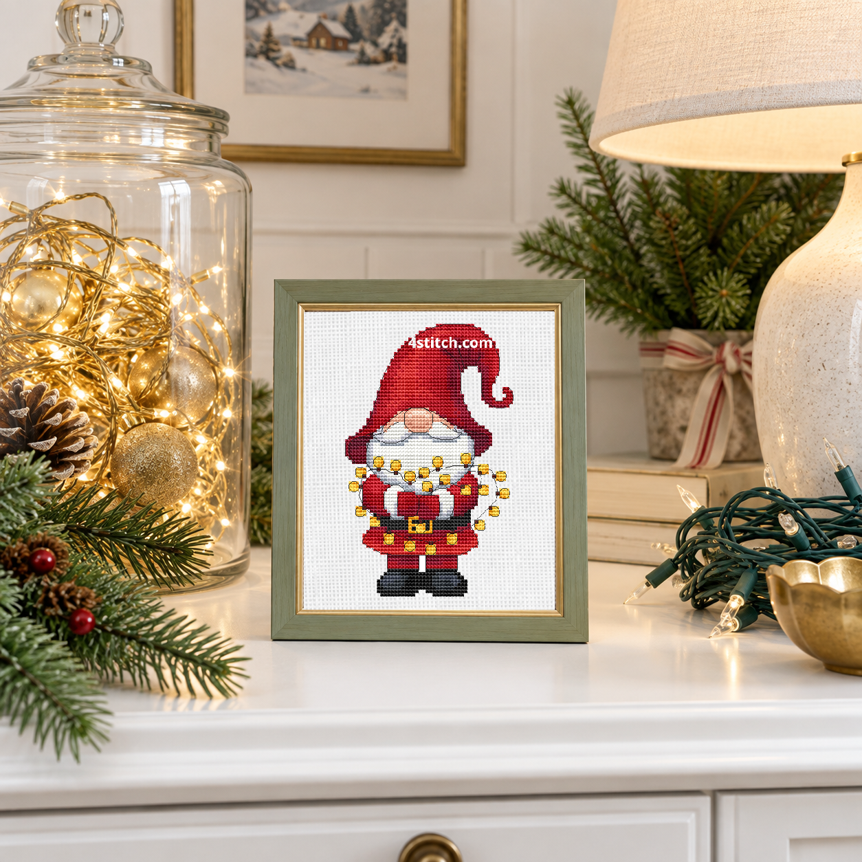 Wider interior view of Christmas Lights Santa Gnome on white Aida in a sage-and-gold frame on a light Christmas decorating console, with the small frame resting among coordinating Christmas decorations.