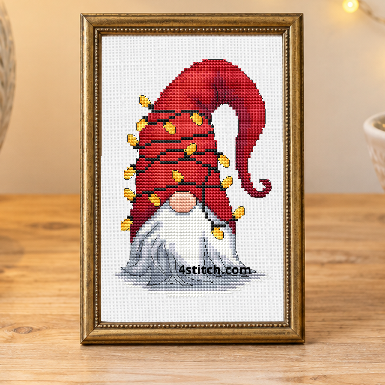 Close-up of Christmas Light Hat Gnome on white Aida in a narrow antique-brass frame in an airy Christmas craft studio.
