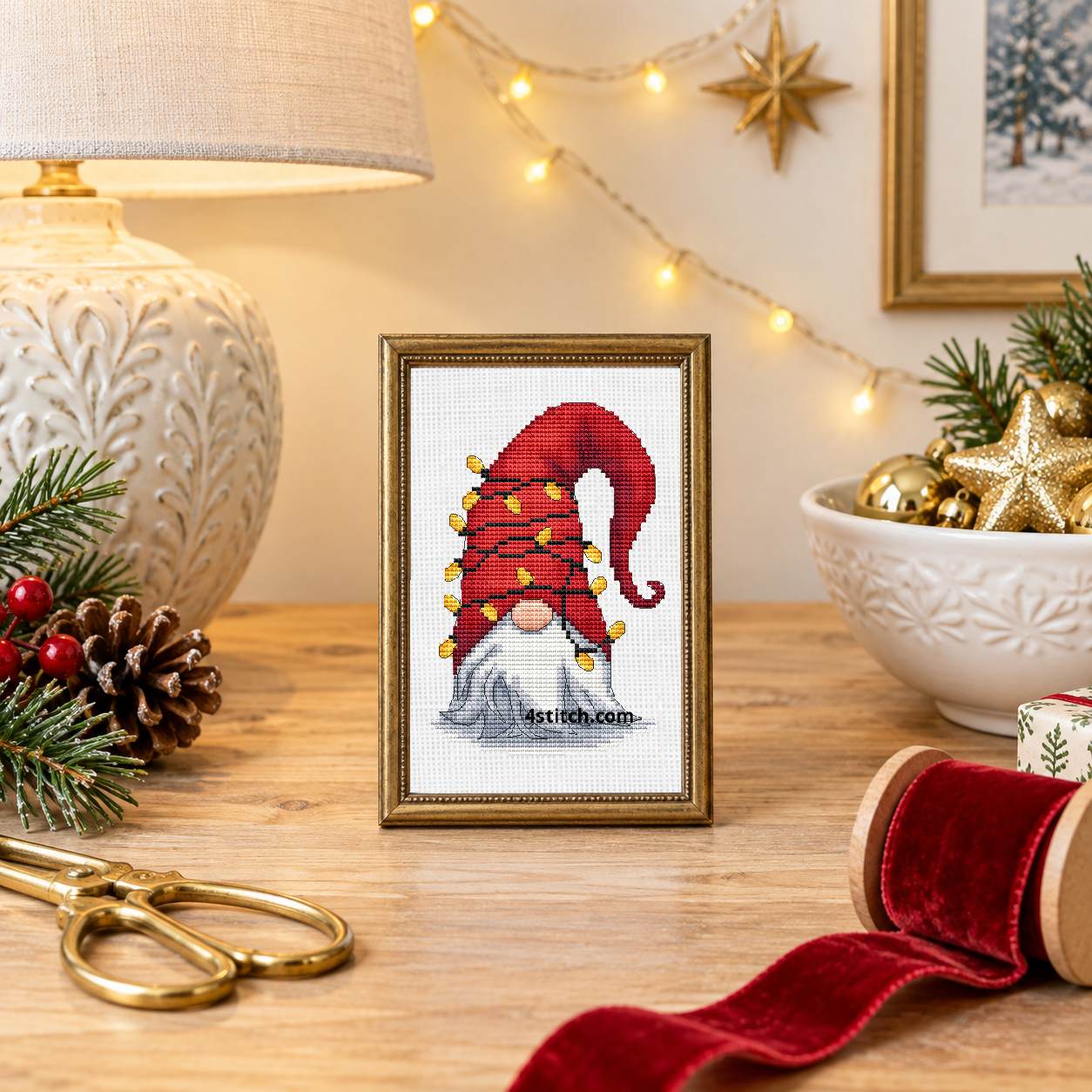 Wider interior view of Christmas Light Hat Gnome on white Aida in a narrow antique-brass frame in an airy Christmas craft studio, with the small frame resting among coordinating Christmas decorations.
