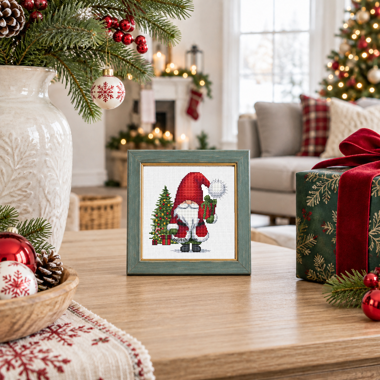 Wider interior view of Christmas Tree Gift Gnome on white Aida in a muted teal frame on a bright festive living-room console, with the small frame resting among coordinating Christmas decorations.