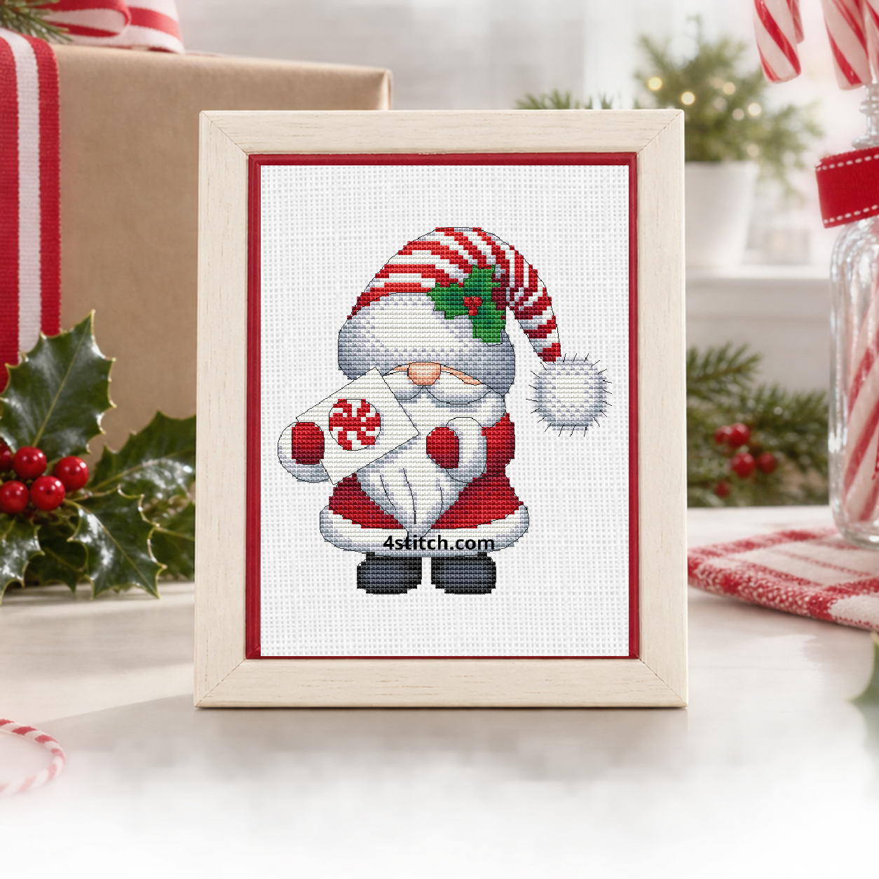 Close-up of Peppermint Gift Santa Gnome on white Aida in an ivory-and-red frame on a bright Christmas wrapping table.