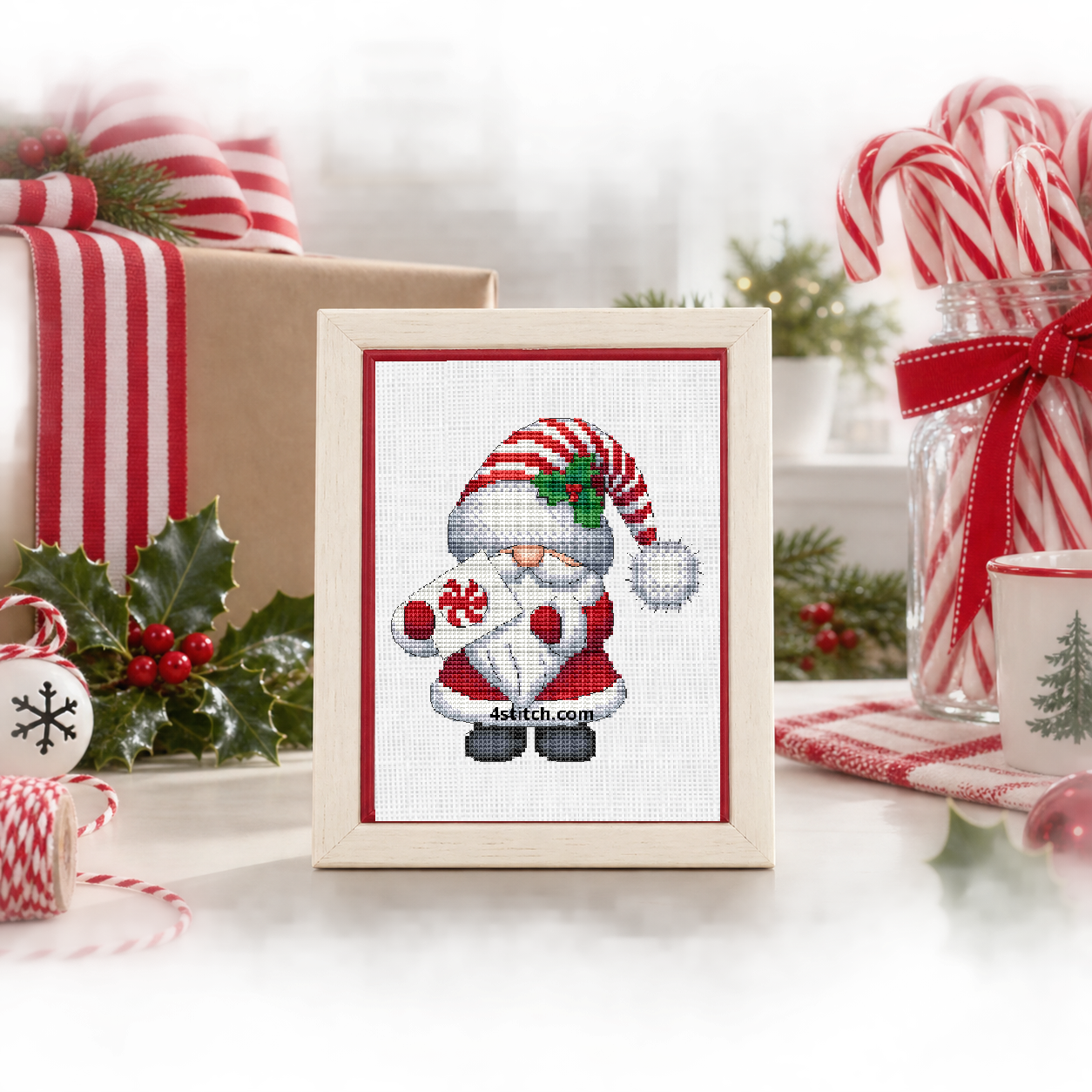 Wider interior view of Peppermint Gift Santa Gnome on white Aida in an ivory-and-red frame on a bright Christmas wrapping table, with the small frame resting among coordinating Christmas decorations.