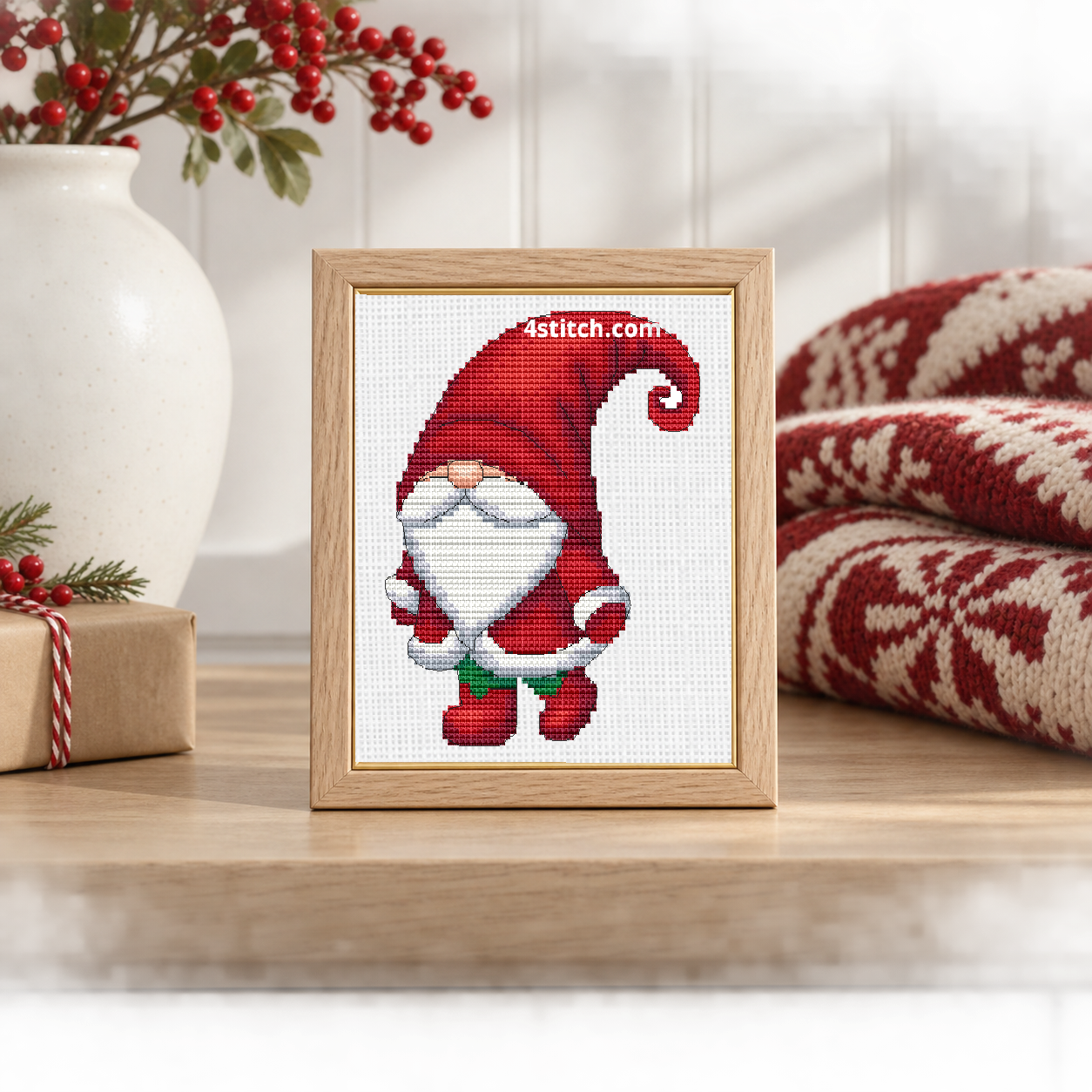 Wider interior view of Curled Hat Santa Gnome on white Aida in a pale oak frame on a light Scandinavian Christmas console, with the small frame resting among coordinating Christmas decorations.
