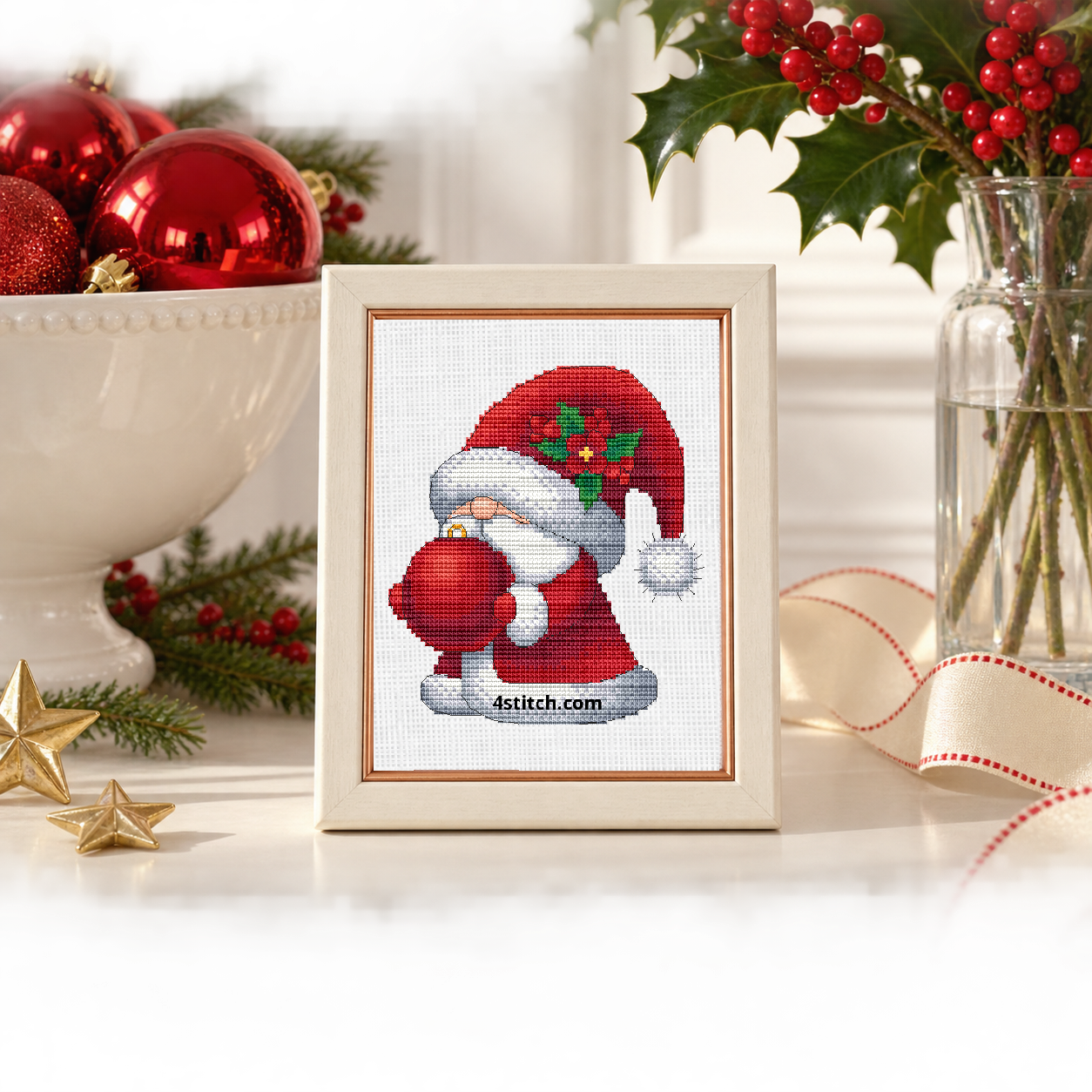 Wider interior view of Holly Bauble Santa Gnome on white Aida in an ivory frame with a copper lip on a bright ornament display, with the small frame resting among coordinating Christmas decorations.