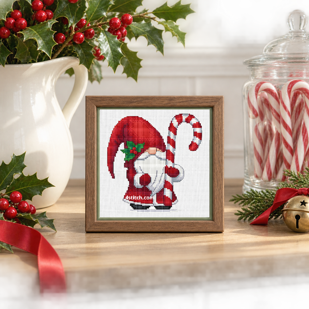 Wider interior view of Holly Candy Cane Gnome on white Aida in a light walnut frame on a cheerful holly-decorated console, with the small frame resting among coordinating Christmas decorations.