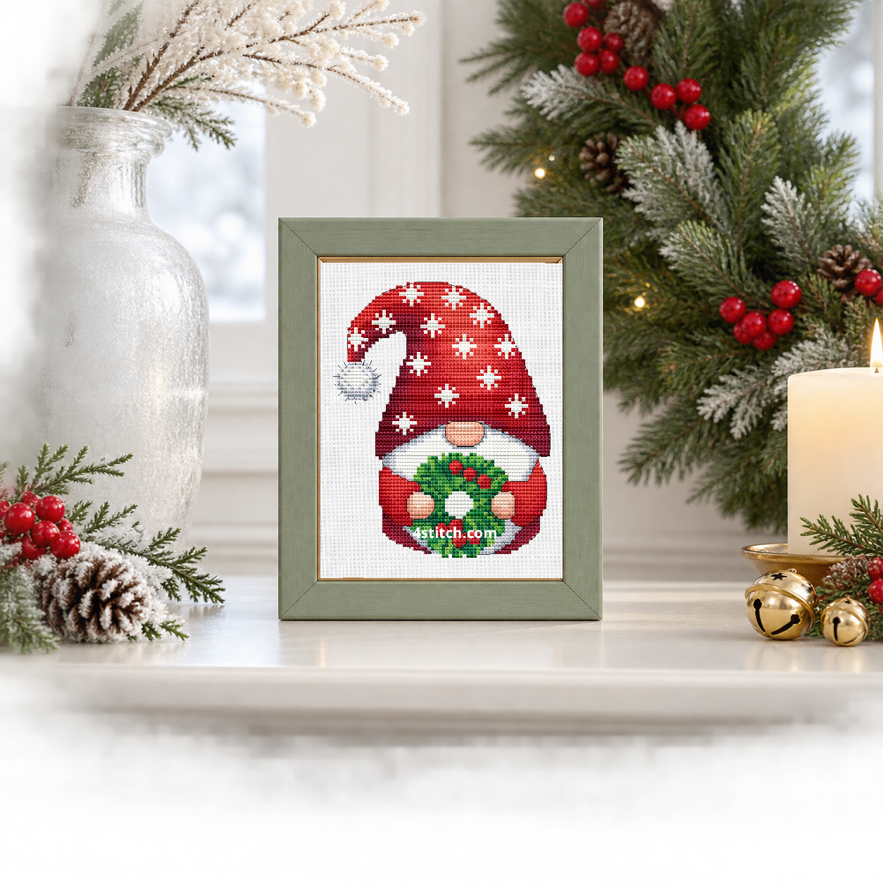 Wider interior view of Snowflake Wreath Christmas Gnome on white Aida in a soft sage-and-gold frame on a luminous winter mantel, with the small frame resting among coordinating Christmas decorations.