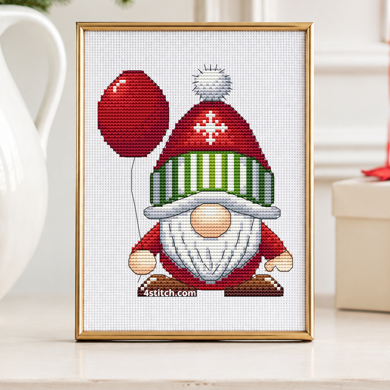 Red Balloon Christmas Gnome with a red balloon, a snowflake hat and a green-and-white striped brim, on white Aida in a champagne brass frame on an ivory console beside winter berries and a red balloon.