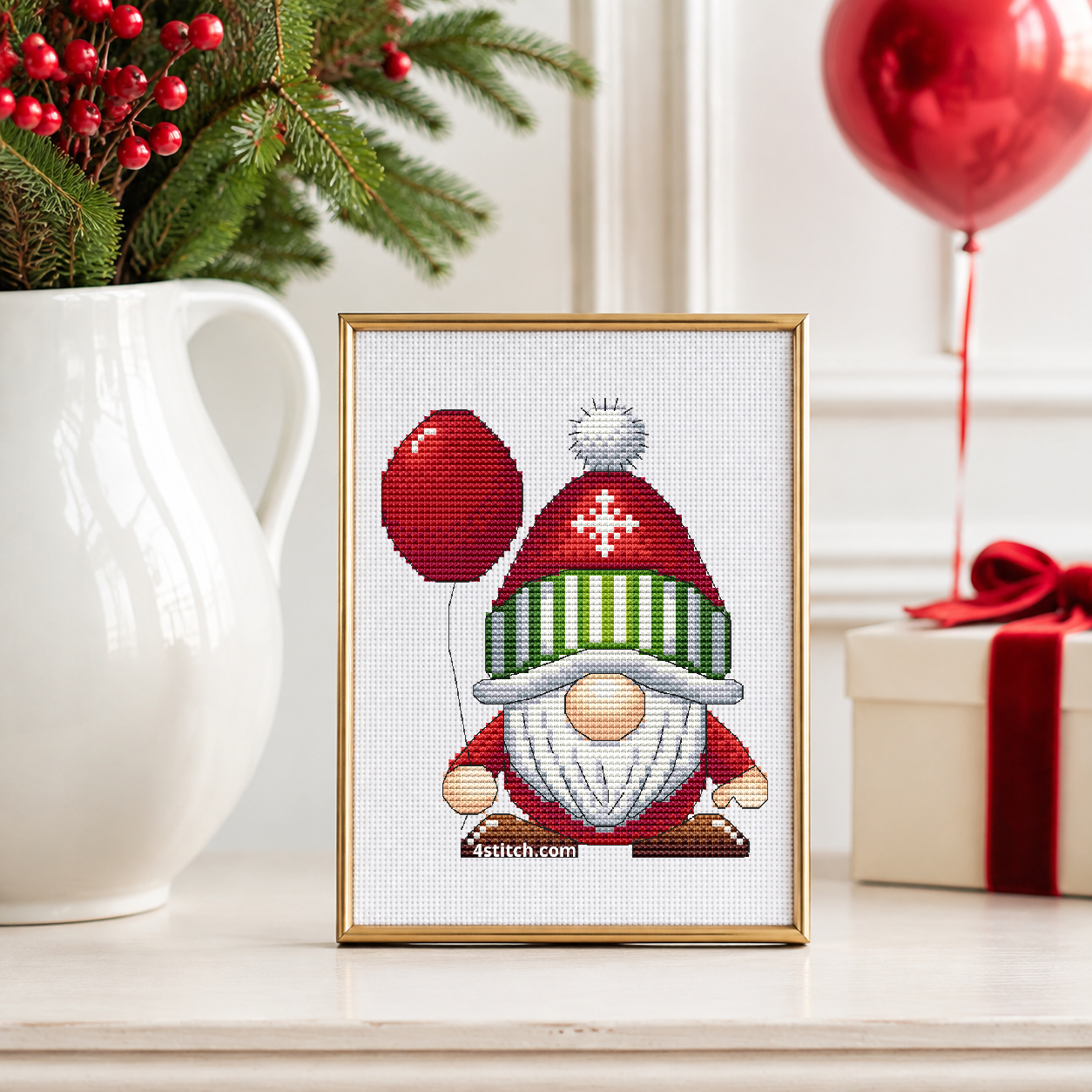 Red Balloon Christmas Gnome with a red balloon, a snowflake hat and a green-and-white striped brim, on white Aida in a champagne brass frame on an ivory console beside winter berries and a red balloon.