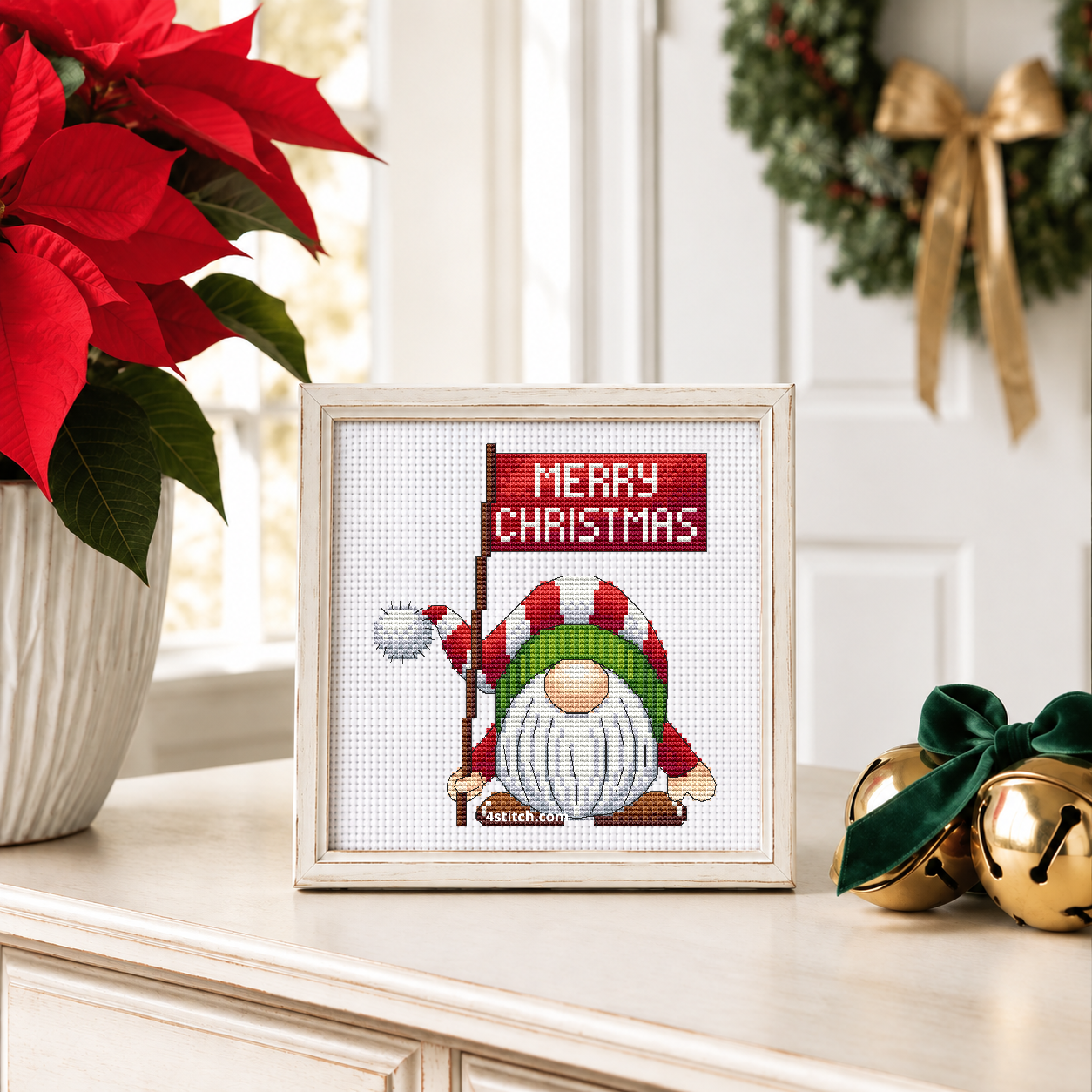 Merry Christmas Sign Gnome with a red Merry Christmas sign and a bent red-and-white striped hat, on white Aida in an ivory frame on a light console with red poinsettia and brass bells.