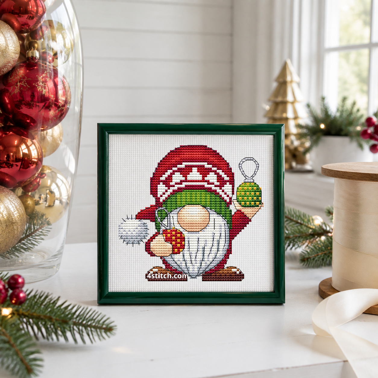 Christmas Ornaments Gnome with red and green hanging baubles, a red patterned hat and a white pompom, on white Aida in a forest-green frame beside a tall glass vase of red and gold ornaments.