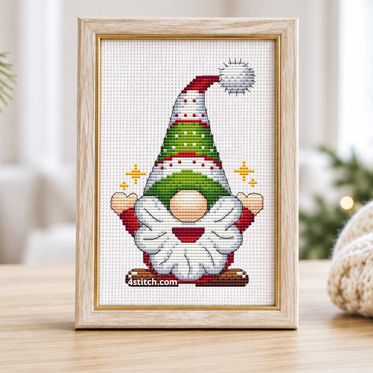 Sparkling Tree Hat Gnome with a tall green-and-red patterned hat and small golden stars beside his hands, on white Aida in a pale birch and gold frame beside pine branches and hanging gold stars.