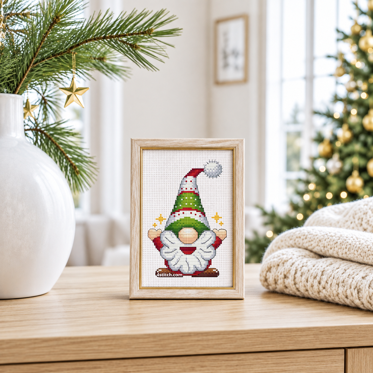 Sparkling Tree Hat Gnome with a tall green-and-red patterned hat and small golden stars beside his hands, on white Aida in a pale birch and gold frame beside pine branches and hanging gold stars.