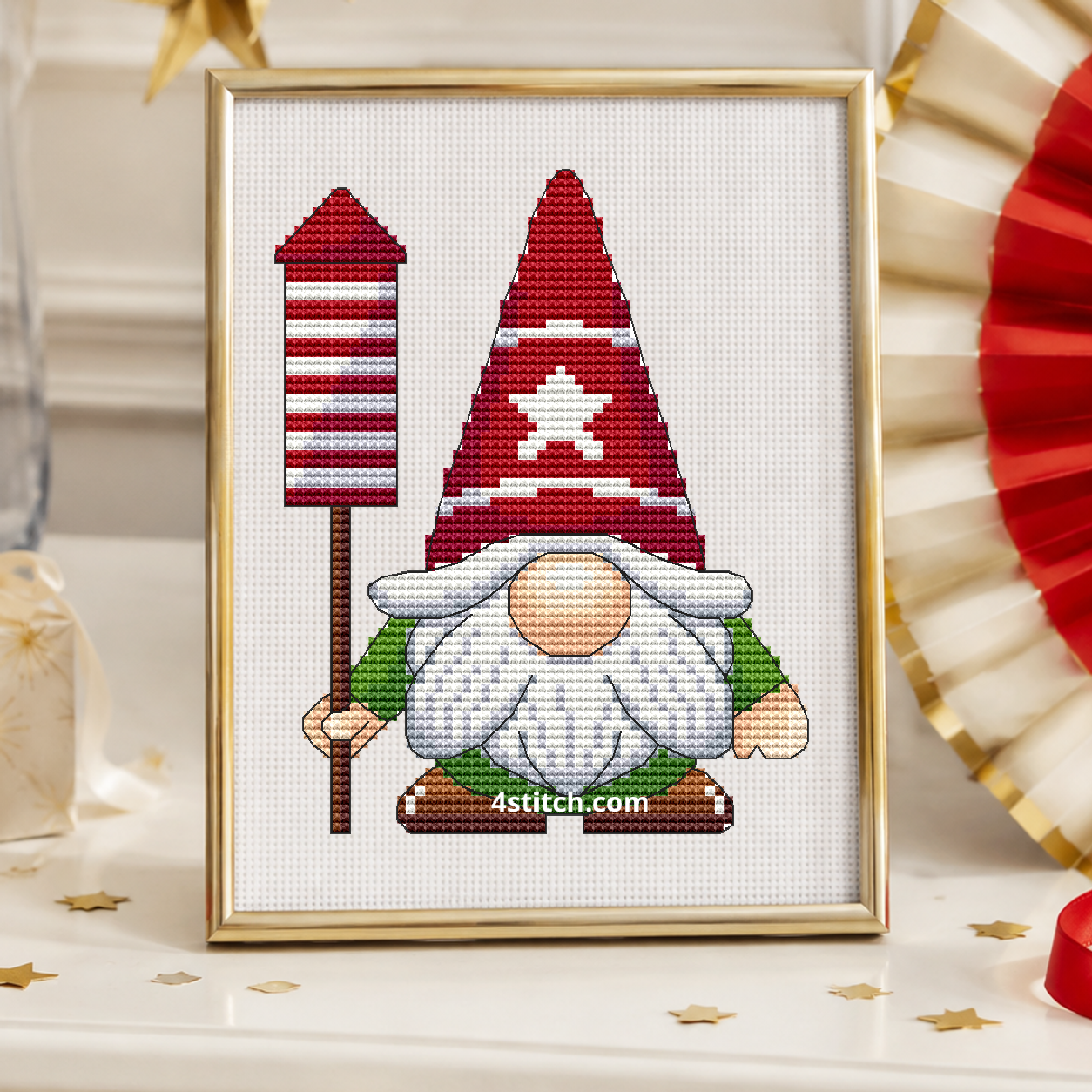 Christmas Rocket Gnome with a red-and-white striped rocket on a pole and a red star-patterned hat, on white Aida in a champagne gold frame on a light holiday console with paper stars and ribbon.