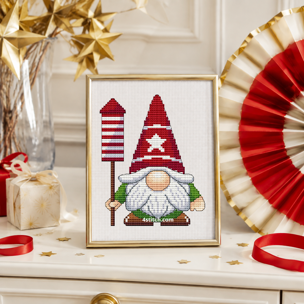 Christmas Rocket Gnome with a red-and-white striped rocket on a pole and a red star-patterned hat, on white Aida in a champagne gold frame on a light holiday console with paper stars and ribbon.