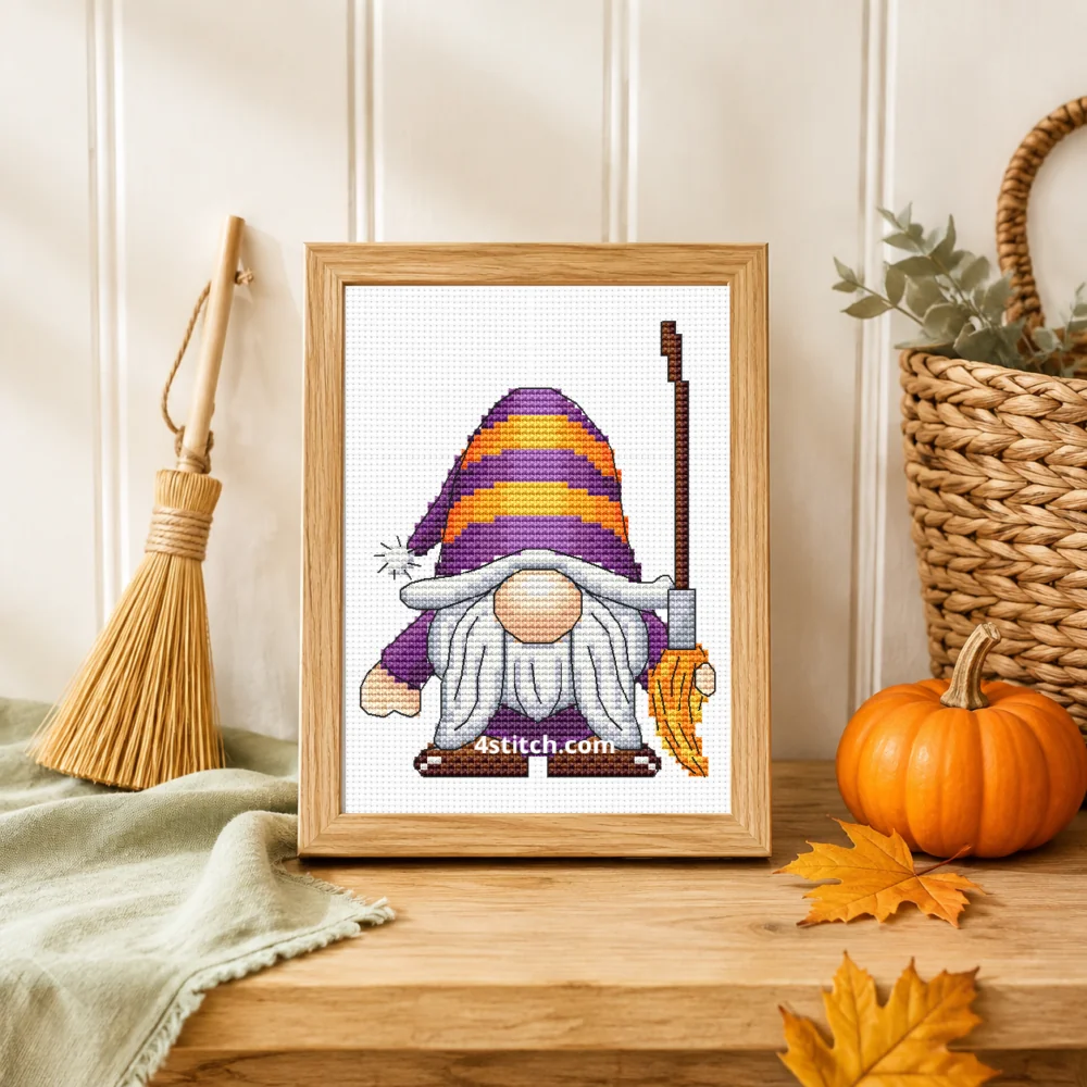 Small framed Striped Hat Broom Halloween Gnome cross stitch on natural white Aida standing on a light table in a bright country interior