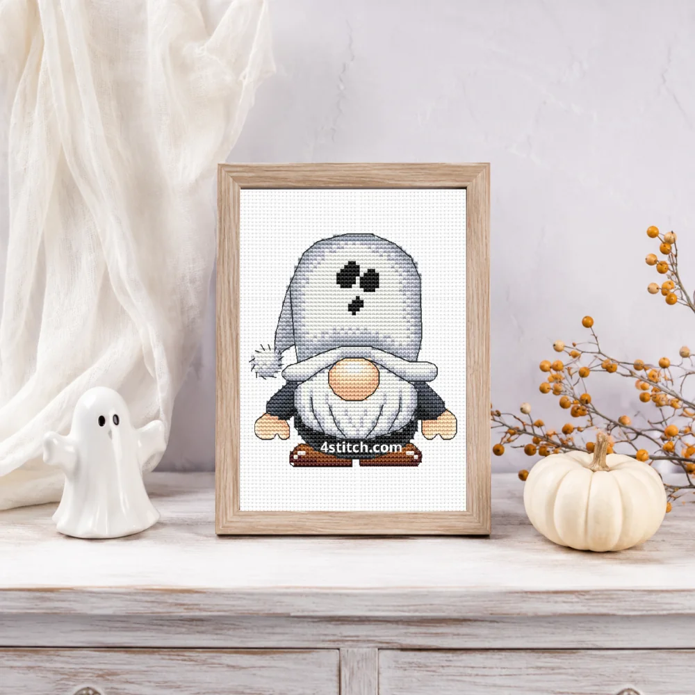 Small framed Ghost Hood Halloween Gnome cross stitch on natural white Aida standing on a light table in a bright ghost-themed interior
