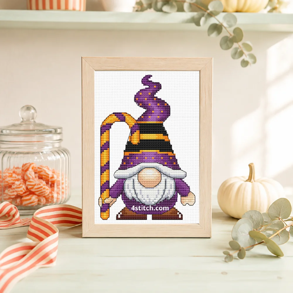 Small framed Candy Cane Witch Halloween Gnome cross stitch on natural white Aida standing on a light table in a bright candy-themed interior